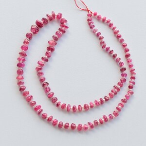 May include: A strand of pink tourmaline beads, with a slightly irregular shape, strung on a red thread. The beads are a light pink color and have a glossy finish.