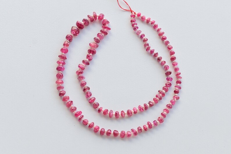 May include: A strand of pink tourmaline beads, with a slightly irregular shape, strung on a red thread. The beads are a light pink color and have a glossy finish.