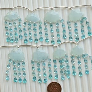 Op de afbeelding: Een zilveren ketting met blauwe edelsteen wolkcharms en blauwe kristallen traanhangers. De ketting is versierd met blauwe kwastcharms.