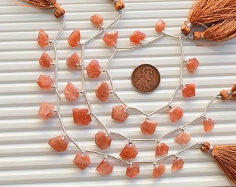 May include: A strand of orange sunstone beads strung on silver wire with tassels on each end. The beads are faceted and have a natural, rough look.