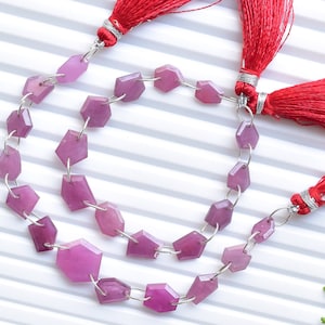 Puede incluir: Una hebra de cuentas de piedras preciosas rosadas con forma hexagonal facetada. Las cuentas están enhebradas en alambre plateado y tienen un borla roja en cada extremo.