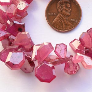 May include: Close-up of a strand of faceted, pink gemstones. The gemstones are irregularly shaped with multiple facets, and are a deep, translucent pink color. A U.S. penny is included for size reference.