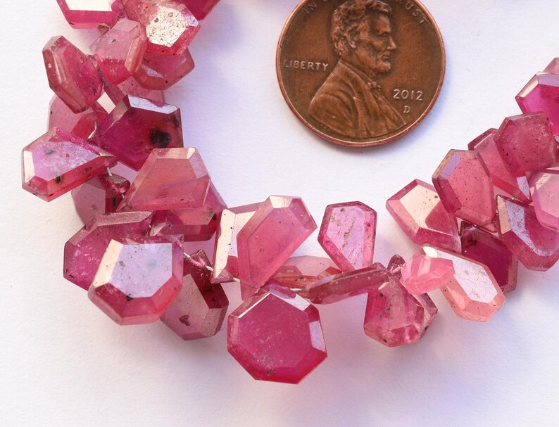 May include: Close-up of a strand of faceted, pink gemstones. The gemstones are irregularly shaped with multiple facets, and are a deep, translucent pink color. A U.S. penny is included for size reference.