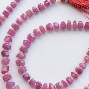 May include: A strand of pink gemstone beads with faceted surfaces. The beads are strung on a white thread and have a slightly irregular shape. The strand is displayed on a white background.
