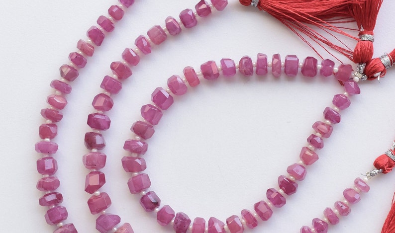 May include: A strand of pink gemstone beads with faceted surfaces. The beads are strung on a white thread and have a slightly irregular shape. The strand is displayed on a white background.