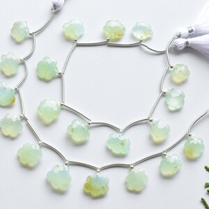 May include: Two strands of pale green flower-shaped beads with silver-colored connecting wires. Each strand has a white tassel at the end. The beads have a translucent quality and a faceted surface. The background is white.