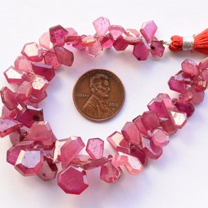 May include: A strand of faceted, pink gemstone beads with a red tassel. The beads are irregularly shaped and vary in size. A US penny is included for size reference. The beads are a deep pink color.