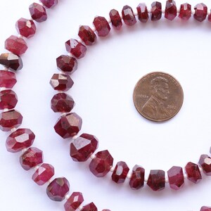 May include: A strand of faceted ruby beads, approximately 8mm in size, with a red colour. The beads are strung on a white thread and are shown next to a UK penny for size comparison.