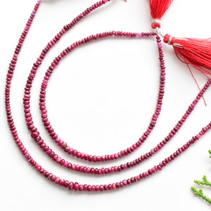 Puede incluir: Tres hebras de cuentas de piedras preciosas de color rojo rubí, cada una con un borla en el extremo. Las cuentas son redondas y lisas, y las borlas están hechas de hilo rojo.