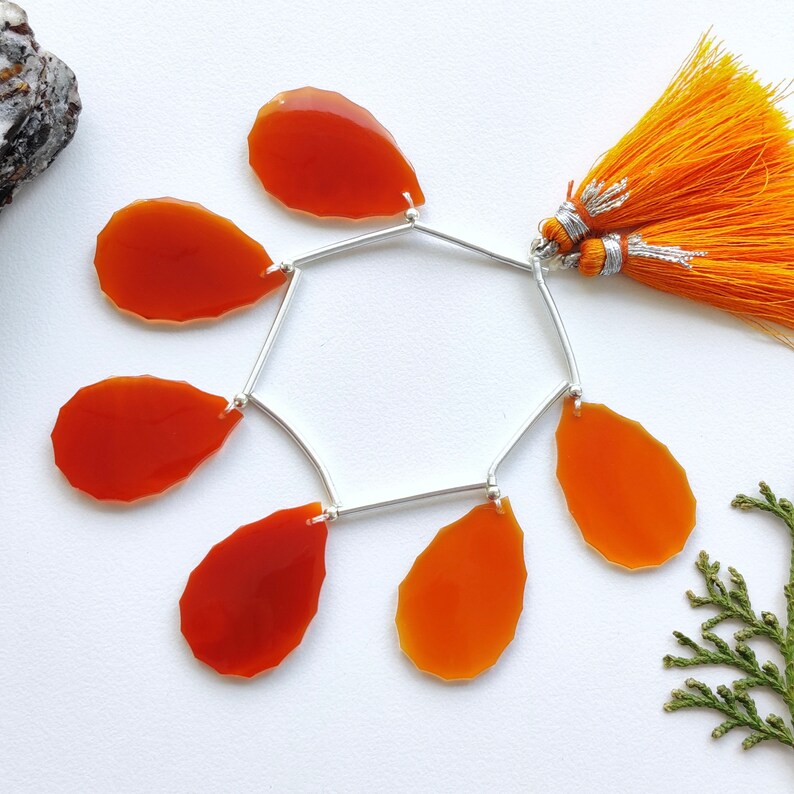 K&ouml;nnte beinhalten: Eine silberne Kette mit sechs orangefarbenen Karneol-Edelsteintropfen und einem doppelten orangefarbenen Quasten. Die Kette hat die Form eines Sechsecks.