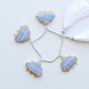 Pode incluir: Uma coleção de pingentes em forma de nuvem de ágata rendada azul com bordas douradas, conectados por um fio verde claro. Dois detalhes de borla branca também estão incluídos, tudo em um fundo branco. Ideal para fazer joias.