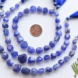 May include: Strands of faceted, blue-violet tanzanite gemstone beads. Each strand is finished with a blue tassel and silver-toned accents. A penny is included for size reference. The gemstones have irregular shapes and a polished finish.
