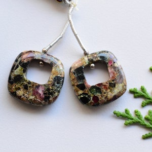 May include: A pair of square-shaped earrings with a square hole in the center. The earrings are made of a dark brown stone with flecks of gold, green, and red. The earrings are hanging from silver chains.