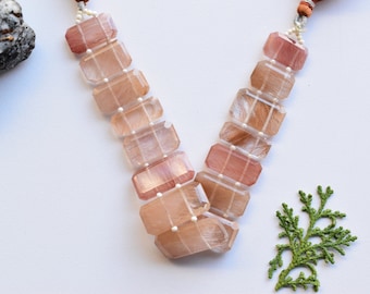 Può includere: Una collana realizzata con pietre rettangolari, traslucide rosa e bianche con un effetto perlescente. Le pietre sono infilate su un cordoncino bianco con piccole perle bianche tra ogni pietra.