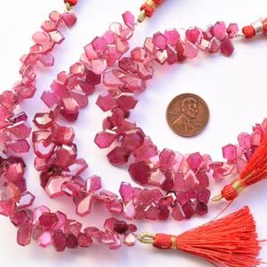 May include: Strands of faceted, pink gemstone beads with a penny for size comparison. The beads are a mix of light and dark pink hues. Red tassels with gold accents are attached to the ends of the strands. The gemstones have an irregular, geometric shape.