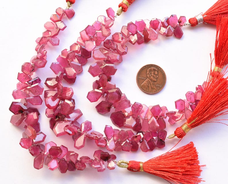 May include: Strands of faceted, pink gemstone beads with a penny for size comparison. The beads are a mix of light and dark pink hues. Red tassels with gold accents are attached to the ends of the strands. The gemstones have an irregular, geometric shape.