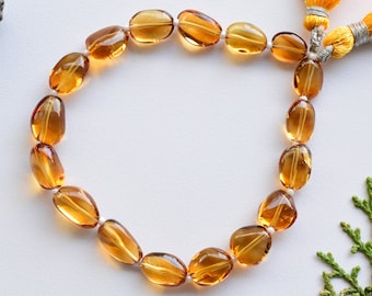May include: A strand of oval-shaped, translucent, amber-colored gemstones strung together with white thread. The strand is laid out in a circle on a white surface.