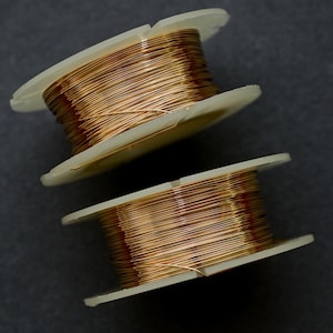 Könnte beinhalten: Zwei Spulen mit Kupferdraht, eine auf der anderen. Die Spulen sind aus weißem Kunststoff und der Draht ist glänzend goldfarben.