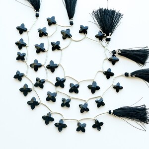 May include: Black gemstone beads in a four-petal flower shape are strung on silver wire, with black tassel accents. The beads are arranged in a circular pattern against a white background, creating a decorative jewelry element.