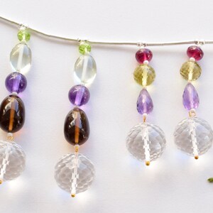 Può includere: Una collana a catena d'argento con più fili di perline di pietre preziose. I fili presentano perline verdi, gialle, viola e rosse. Le perline sono sfaccettate e hanno un aspetto chiaro e cristallino.