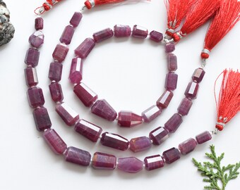 May include: Strands of faceted, deep red ruby beads. Each bead is a rectangular shape with multiple facets, strung together with silver-colored string. Red tassels are attached to the ends of the strands. The beads are displayed on a white surface.