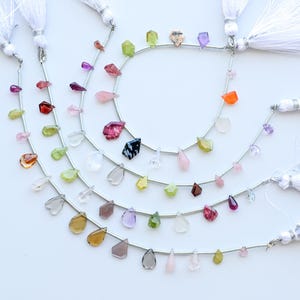 May include: A selection of gemstone bead strands featuring a variety of shapes and colours, such as teardrops and faceted stones. The beads are strung on silver wire and finished with white tassel ends. Gemstones include red, pink, green, yellow, and purple.