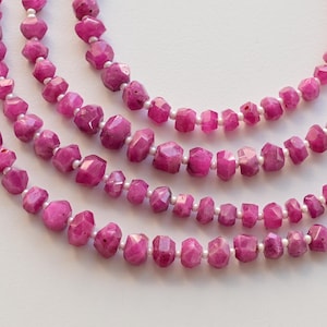 以下が含まれることがあります： 白い糸に、ファセットカットされたピンクのルビービーズが連なっています。ビーズは重ねて配置され、繊細でエレガントな外観になっています。