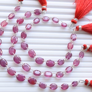 Puede incluir: Una hebra de cuentas de piedras preciosas ovaladas de color rosa con conectores plateados y borlas rojas.