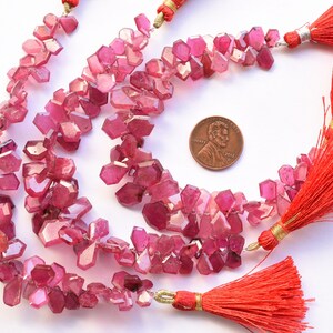 May include: Strands of faceted, pink gemstone beads with a penny for size comparison. The beads are clustered and irregular in shape. Bright red tassels with gold accents are attached to the strands.