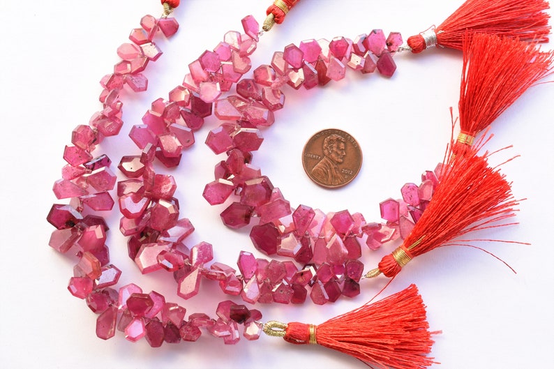 May include: Strands of faceted, pink gemstone beads with a penny for size comparison. The beads are clustered and irregular in shape. Bright red tassels with gold accents are attached to the strands.