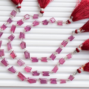 Puede incluir: Una hebra de cuentas de piedras preciosas rosadas con forma rectangular facetada. Las cuentas están enhebradas en alambre plateado y tienen un acabado pulido. La hebra está adornada con borlas rojas.