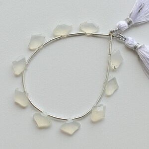 May include: A strand of pale, faceted gemstone beads with a silver-colored wire, and white tassels. The beads are irregularly shaped and translucent. The tassels are made of white threads, and are attached to the wire.