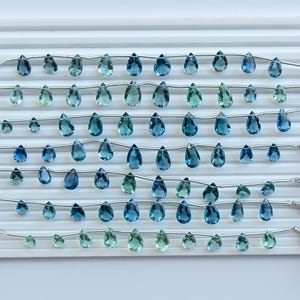 May include: A display of blue and teal teardrop gemstones on a white tray. Each strand features multiple gems of varying sizes. White tassels adorn the ends. The gemstones are polished and translucent. The arrangement suggests a jewellery-making theme.
