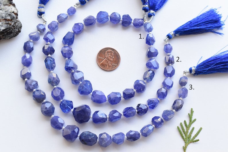 May include: Three strands of faceted, blue-violet tanzanite gemstone beads. Each strand is finished with a blue tassel and silver-toned accents. A U.S. penny is included for size reference. The beads have an irregular, geometric shape.