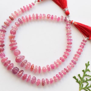 Könnte beinhalten: Eine Kette aus rosafarbenen Edelsteinperlen mit je einem roten Quasten an jedem Ende. Die Perlen sind oval geformt und haben eine leicht unregelmäßige Form. Die Perlen sind auf einem Silberdraht aufgefädelt.