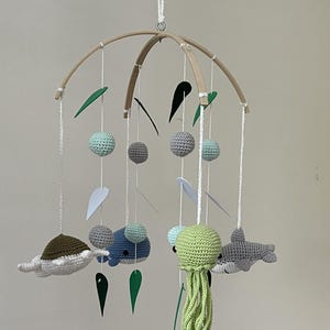 May include: A wooden baby mobile with crocheted sea creatures, including a turtle, whale, jellyfish, and shark. The mobile also features green felt leaves and gray and blue felt balls.