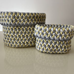 May include: Two handmade, knitted baskets in a neutral color palette. The baskets are in a mix of yellow, gray, and white. The larger basket is approximately 6 inches in diameter and 5 inches tall, while the smaller one is about 4 inches in diameter and 3 inches tall.