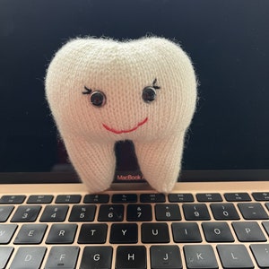 Könnte beinhalten: Ein weißer gestrickter Zahn mit schwarzen Augen und einem roten Lächeln, der auf einer Laptop-Tastatur sitzt.