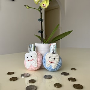 Può includere: Due portafogli a forma di dente in crochet, uno rosa e uno blu, con facce sorridenti. I portafogli sono su un tavolo bianco con una piccola pianta di orchidea in vaso dietro di loro. Ci sono diverse monete sparse intorno ai portafogli.