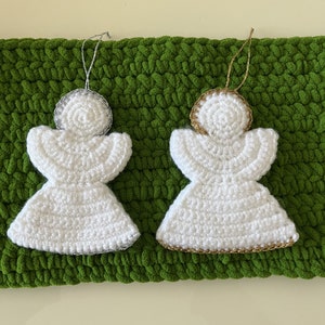May include: Two white crocheted angel ornaments with gold and silver trim. The ornaments are hanging from a green crocheted surface.