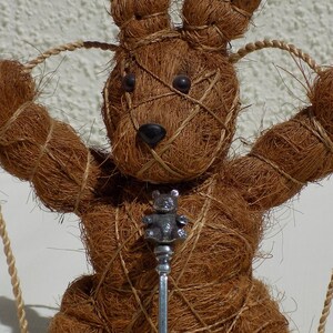 Peut inclure: Un lapin en fibre de noix de coco brun assis sur une balançoire avec une cuillère en argent suspendue à son cou.