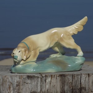 May include: A ceramic figurine of a dog, painted in shades of beige and brown, with a teal collar, standing on a green base. The dog is positioned as if it is sniffing the ground.