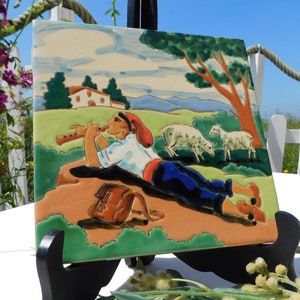 Può includere: Una piastrella in ceramica che raffigura un pastore che suona il flauto mentre giace su una collina con pecore sullo sfondo. La piastrella è dipinta in tonalità di verde, marrone, blu e rosso.