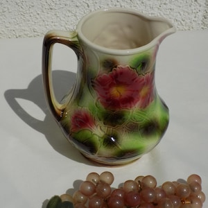 Puede incluir: Una jarra de cerámica vintage con un diseño floral en tonos verdes, marrones y rojos. La jarra tiene un asa y una boca ancha. Un racimo de uvas artificiales con hojas verdes está en primer plano.