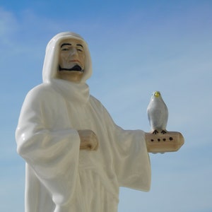 Op de afbeelding: Een wit porseleinen beeldje van een man die een witte robe draagt en een witte vogel in zijn rechterhand vasthoudt. De man heeft een baard en snor en kijkt naar rechts. De achtergrond is een blauwe lucht.