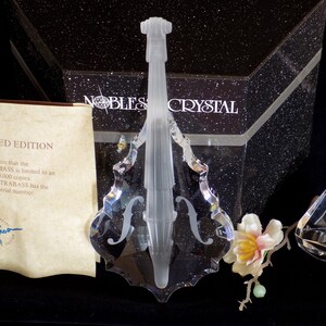 Op de afbeelding: Een transparant kristallen beeld van een contrabas, met een bloem en een kleiner kristallen beeld op de achtergrond. Het beeld is onderdeel van een gelimiteerde editie van 3.000 stuks. De tekst "NOBLES CRYSTAL" is zichtbaar op de doos.