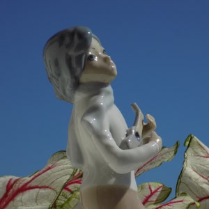 Puede incluir: Una figurita de porcelana de una persona joven con ojos azules y un vestido blanco, sosteniendo un pequeño animal de porcelana. La figurita está colocada sobre un fondo de cielo azul y hojas blancas y rojas.