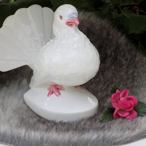 May include: White ceramic dove figurine with a pink base and pink feet. The dove has its tail feathers fanned out and is standing on a small oval base. A pink rose is in the foreground.