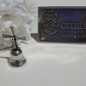 May include: A silver bell with a windmill design on top. The bell is sitting on a white surface. A silver plaque with the text "GELRIA TIN" and "waarborg voor kwaliteit" is in the background.