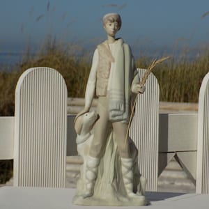 May include: A porcelain figurine of a young shepherd with a dog, holding a staff with wheat stalks. The shepherd is wearing a brown vest and a long, light blue scarf. The figurine is standing on a base with a green and brown design.